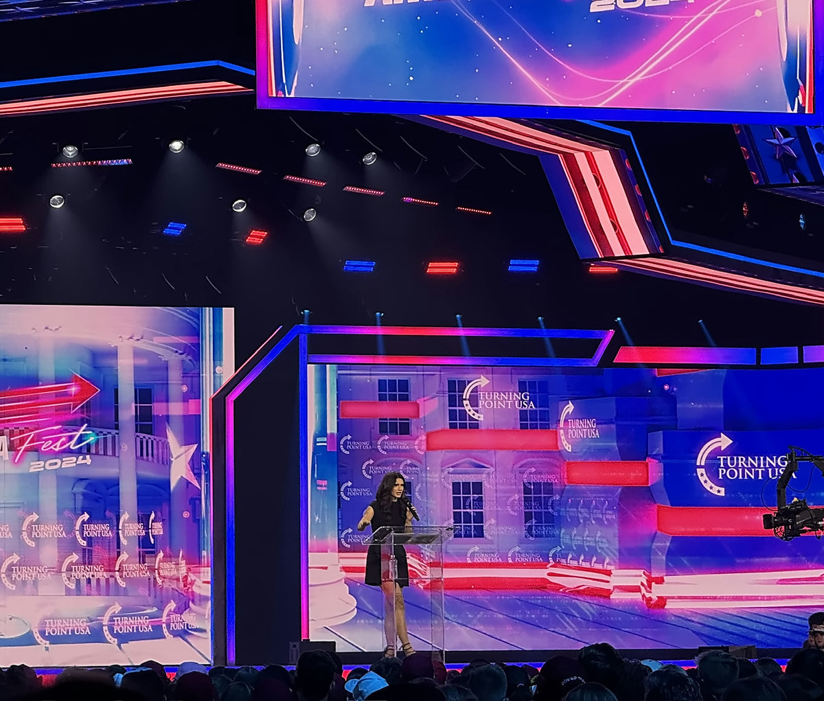 Speaker at a clear podium on a large event stage with LED screens, red and blue lighting and a silhouetted crowd