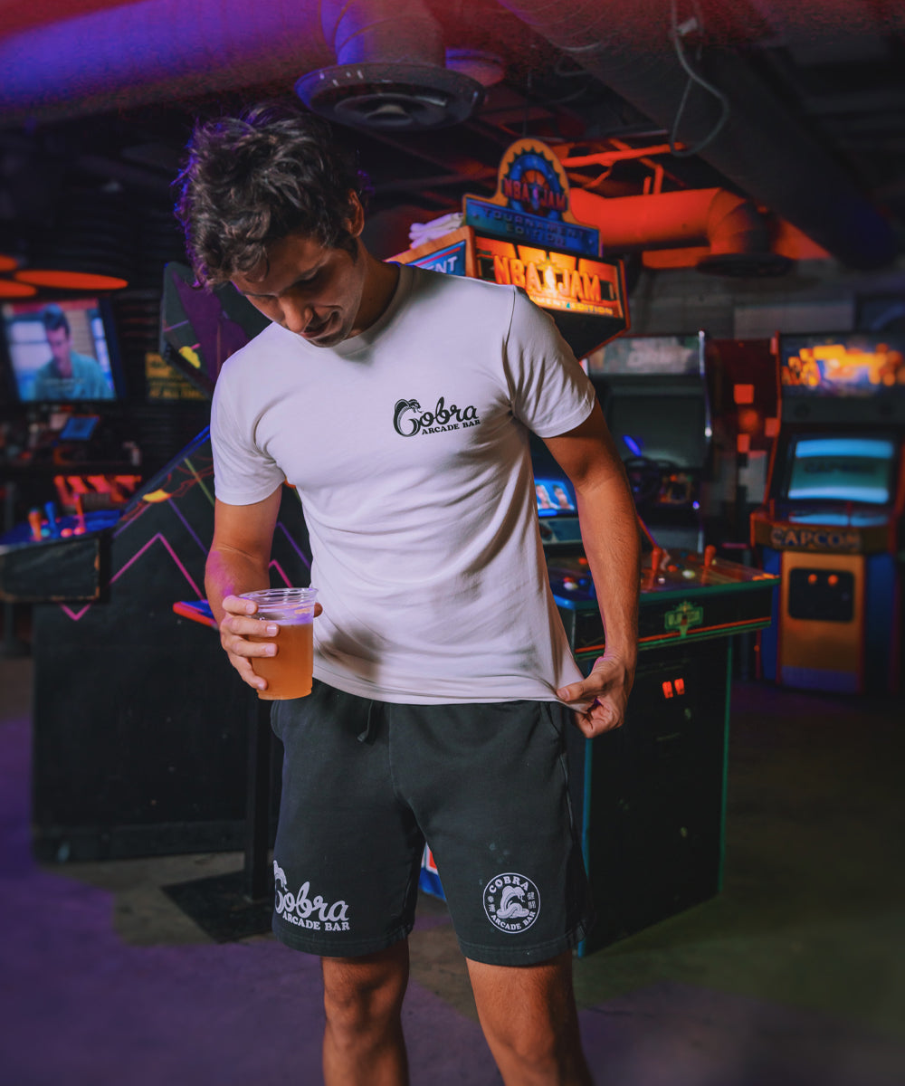 Man in a white tee and black shorts branded Cobra Arcade Bar, standing in an arcade
