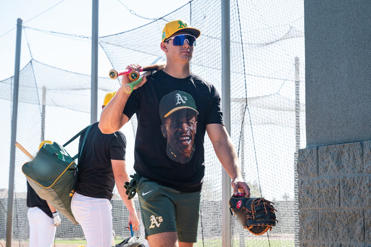 A player in the Rickey Henderson tribute tee carrying bats past the batting cages, with a teammate in the same tee behind him