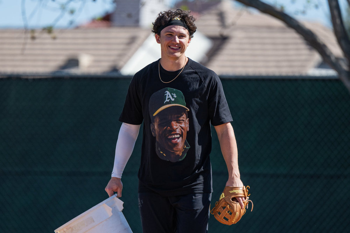 A smiling player in the Rickey Henderson tribute tee carrying a glove and a bucket