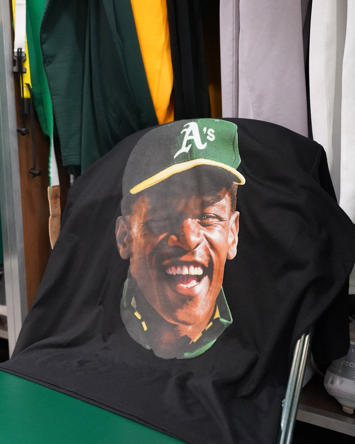 Rickey Henderson tribute tee draped over a green chair in the A's clubhouse, printed by Superluxe Merch