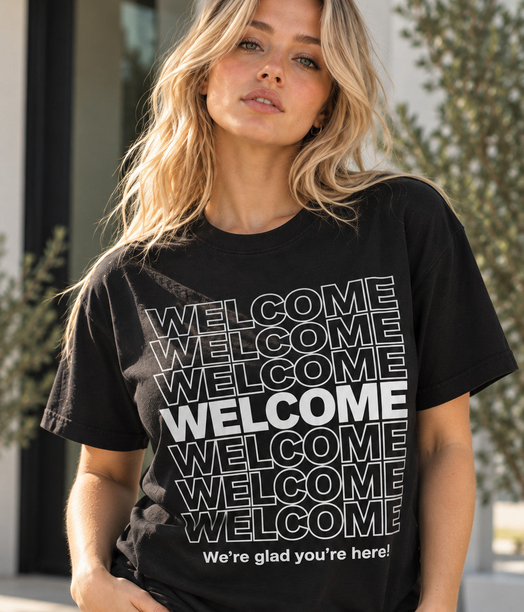 Woman outdoors in daylight wearing a black custom tee printed with the word WELCOME stacked seven times and "We're glad you're here!" underneath
